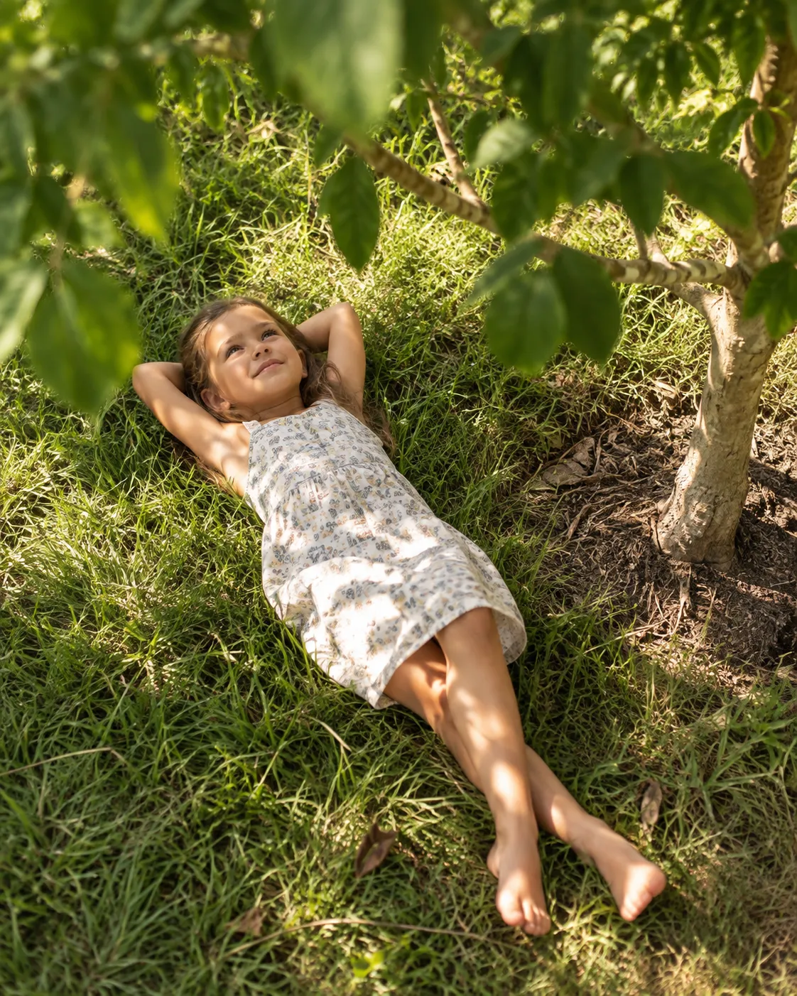 A child lying barefoot in sunlit grass, looking up through the leaves of a young tree